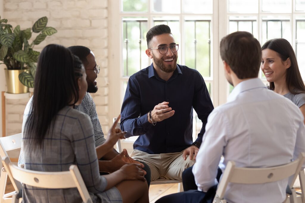 A diverse group of professionals sit in a circle during a workplace discussion as one man speaks and others listen.